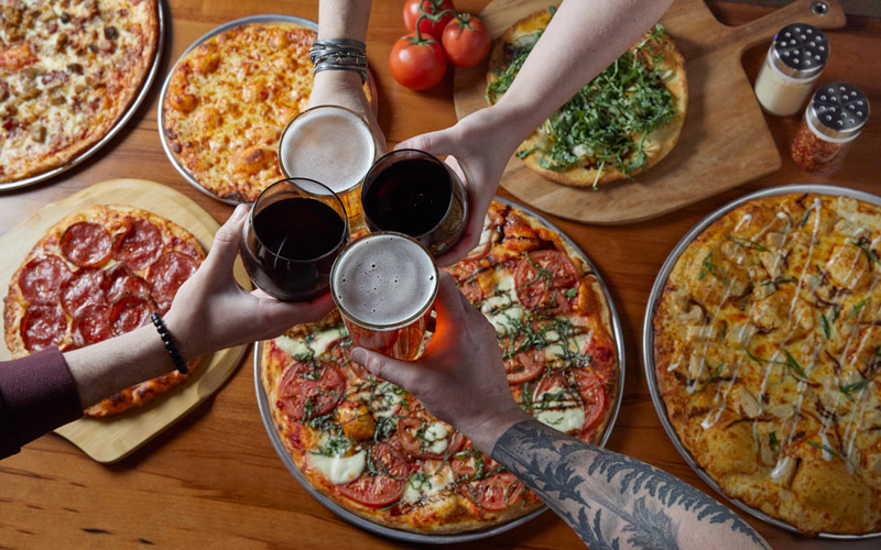 A picture of four hands holding beverages, clinking them together in "Cheers" with an assortment of pizzas in the background, from the restaurant at Arena Sports