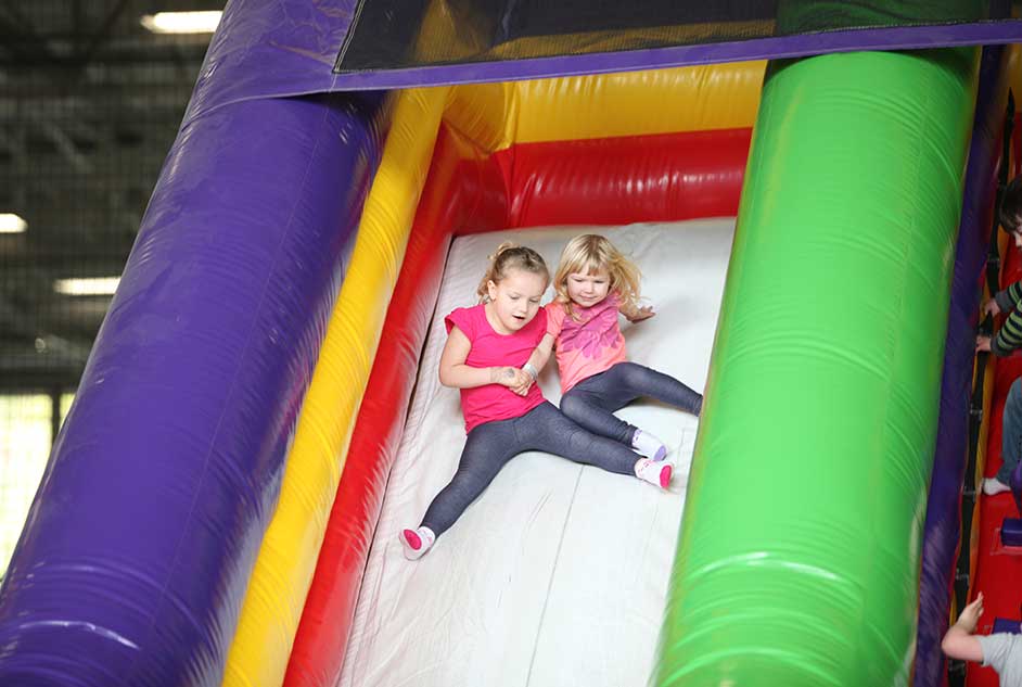 Two young sisters slide down a bouncy house slide in the Inflatable FunZone indoor playground at Arena Sports