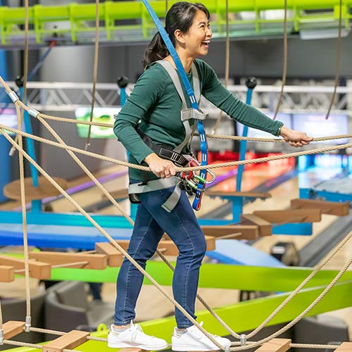 A woman wearing a safety harness and holding the rope lines smiles while walking the ropes course at Arena Sports Mill Creek